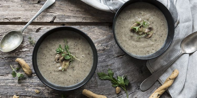 due ciotole scure con la vellutata di funghi, cucchiai e grissini sparsi su tavolo di legno