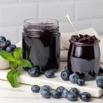 In primo piano due vasetti di vetro con all'interno la marmellata di mirtilli neri e dei mirtilli sul vassoio