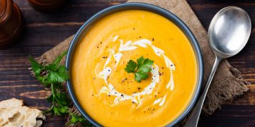 zuppe vellutate: su base di legno scodella scura piena di vellutata di zucca, prezzemolo, un cucchiaio e del pane accanto