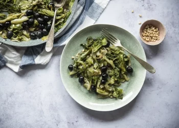 su piano di marmo, piatto verde con della scarola stufata con pinoli, accanto un contenitore piccolo con dei pinoli e dietro un canovaccio con un piatto di scarola