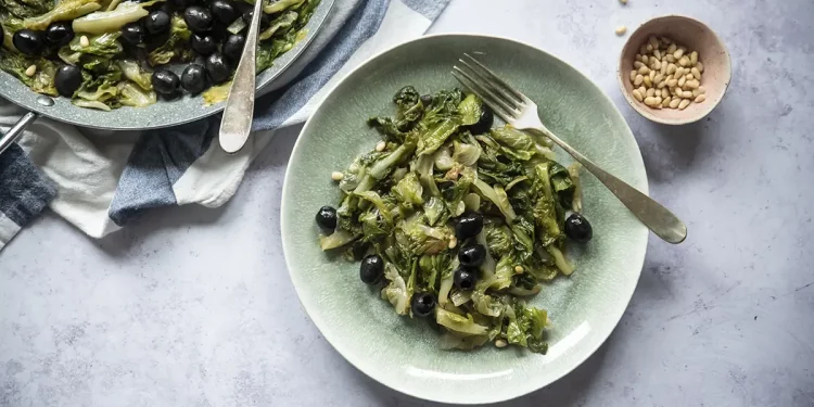 su piano di marmo, piatto verde con della scarola stufata con pinoli, accanto un contenitore piccolo con dei pinoli e dietro un canovaccio con un piatto di scarola