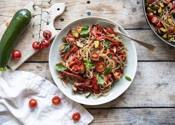 su tavolo di legno piatto con shirataki di konjac con verdure, accanto canovaccio con pomodorini e sul tagliere di legno zucchine . A destra padella con spaghetti di konjac