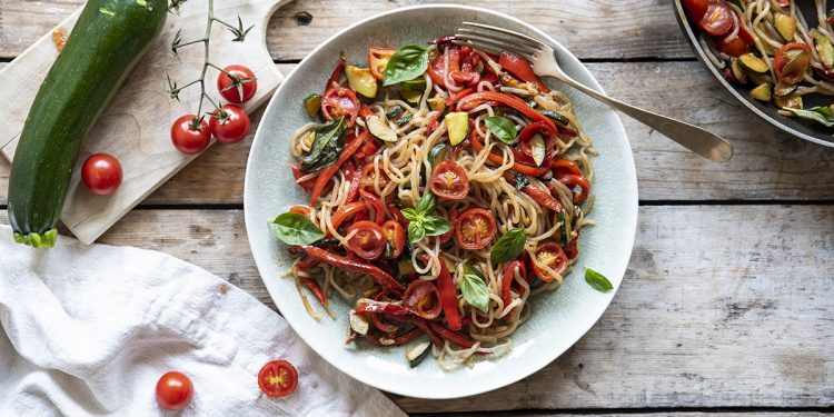 su tavolo di legno piatto con shirataki di konjac con verdure, accanto canovaccio con pomodorini e sul tagliere di legno zucchine . A destra padella con spaghetti di konjac