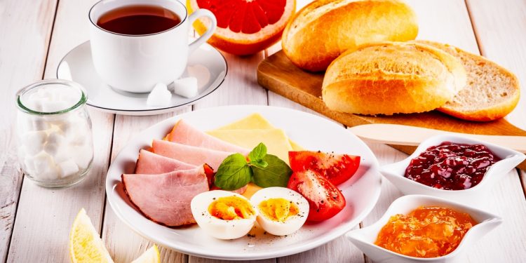 colazione proteica: un piatto con uova e prosciutto cotto, una tazza di caffè, pane, arancia e marmellate