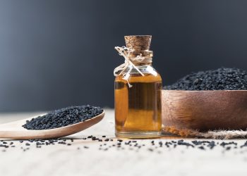 una boccetta di vetro con tappo di sughero piena di olio e intorno ciotola e cucchiaio di legno con semi di cumino