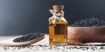 una boccetta di vetro con tappo di sughero piena di olio e intorno ciotola e cucchiaio di legno con semi di cumino