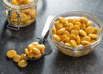 su tavolo nero, un contenitore di vetro pieno di lupini, un cucchiaio di lupini e dietro un barratolo pieno di semi di lupino