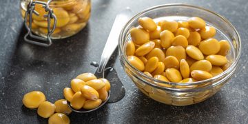 su tavolo nero, un contenitore di vetro pieno di lupini, un cucchiaio di lupini e dietro un barratolo pieno di semi di lupino