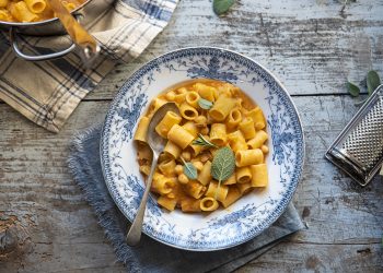 Su tavolo di legno piatto colorato con pasta e ceci e un foglio di alloro, accanto grattugia e il parmigiano. In alto a sinistra la padelle con pasta e ceci