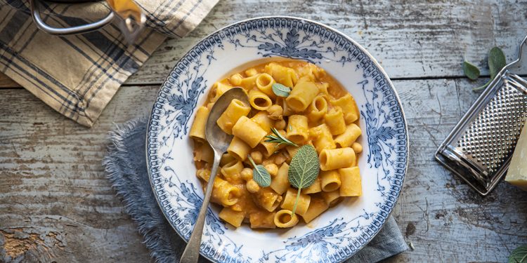 Su tavolo di legno piatto colorato con pasta e ceci e un foglio di alloro, accanto grattugia e il parmigiano. In alto a sinistra la padelle con pasta e ceci