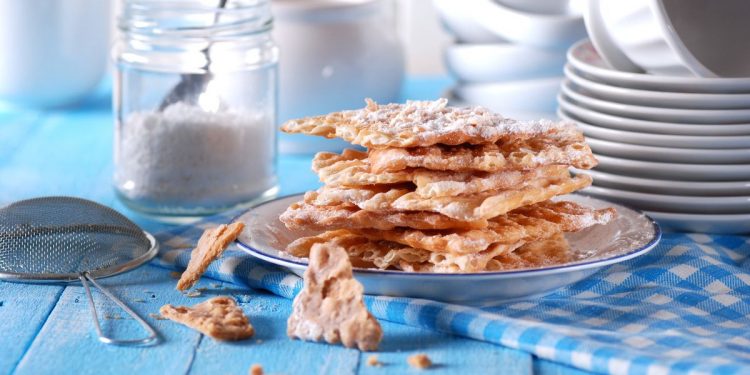 Dolci di carnevale: su tavola azzurra un piatto con le chiacchiere al forno e un vasetto di zucchero accanto