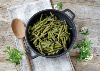 Ricette con fagiolini: padella con manici nera piena di fagiolini verdi, un cucchiaio e erbette accanto
