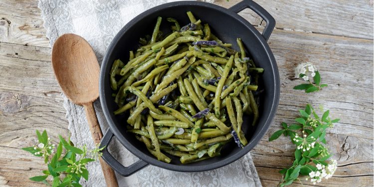 Ricette con fagiolini: padella con manici nera piena di fagiolini verdi, un cucchiaio e erbette accanto