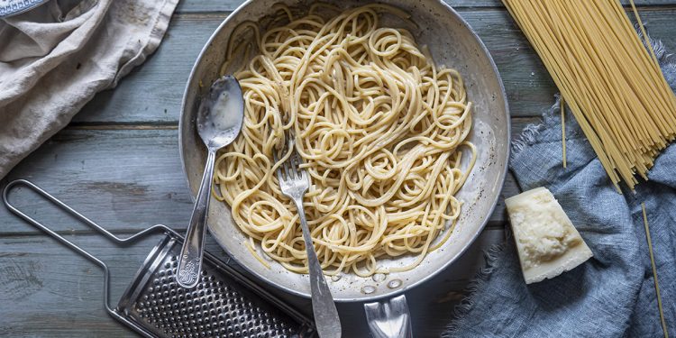 su tavolo di legno, in un padella spaghetti cacio e pepe, con accanto la grattugia, il Pecorino e gli spaghetti