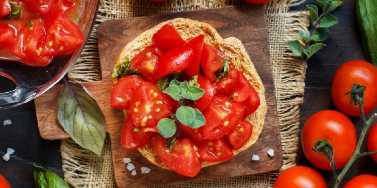 ricette con pomodori: su piatto di legno una frisella con pomodorini e foglie di basilico