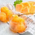 Granita al melone: la ricetta golosa senza gelatiera 1 In primo piano su uno sfondo chiaro ci sono due coppette in vetro con la granita al melone, un cucchiaino dorato e sullo sfondo delle fette di melone fresco.