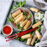 vassoio rettangolare con tanti involtini primavera, alcuni in un piatto tondo, ciotola salsa agrodolce e bacchette cinesi