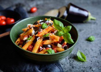 su tavolo di legno, ciotola verde con pasta alla Norma e attorno ingredienti, pomodori, melanzane e basilico