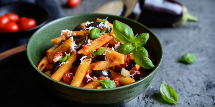 su tavolo di legno, ciotola verde con pasta alla Norma e attorno ingredienti, pomodori, melanzane e basilico