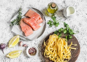 au base chiara un piarro con tre tranci di salmone crudo, un piatto con delle tagliatelle crude, boccetta di olio, spicchi di limone e prezzemolo