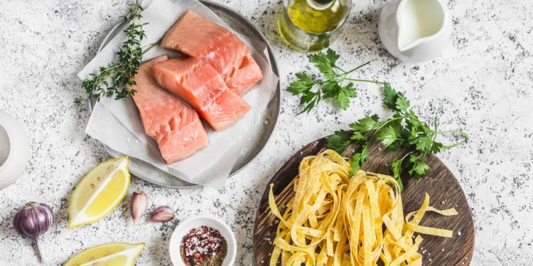 au base chiara un piarro con tre tranci di salmone crudo, un piatto con delle tagliatelle crude, boccetta di olio, spicchi di limone e prezzemolo