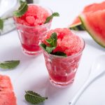 Sorbetto all’anguria fresco e dissetante 1 In primo piano su uno sfondo bianco, ci sono due bicchieri di vetro con del sorbetto all'anguria decorati da delle foglioline di menta.