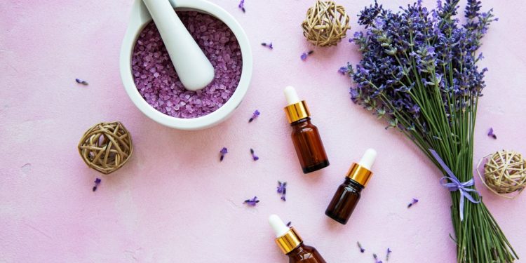 su base lilla un mortaio bianco con foglie di lavanda, un mazzetto di lavanda e 3 flaconcini