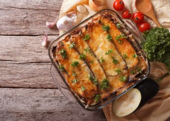 Su tavolo di legno, pirofila con la moussaka, aglio, pomodorini e erbe aromatiche