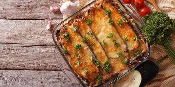 Su tavolo di legno, pirofila con la moussaka, aglio, pomodorini e erbe aromatiche