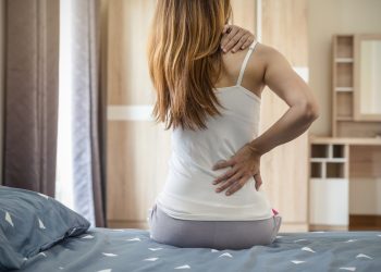 dormire con il mal di schiena: una giovane donna che si alza dal letto tenendosi la schiena con la mano