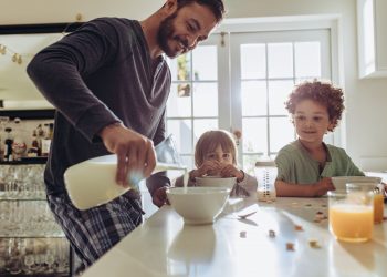 un papà che versa del latte ai due figli per colazione