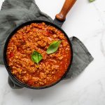 In primo piano su uno sfondo di marmo chiaro una pentola nera con il ragù alla bolognese, decorato con una foglia di basilico.