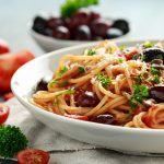 Pasta alla puttanesca: ricetta veloce e saporita 1 su tavolo primo piano di un piatto con spaghetti alla puttanesca, una ciotola con olive nere, aglio e pomodorini tagliati