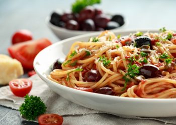 su tavolo primo piano di un piatto con spaghetti alla puttanesca, una ciotola con olive nere, aglio e pomodorini tagliati