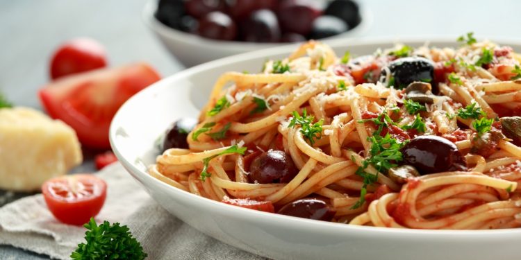 su tavolo primo piano di un piatto con spaghetti alla puttanesca, una ciotola con olive nere, aglio e pomodorini tagliati