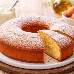 In primo piano su un tavolo di legno rustico c'è un piatto fondo con la torta 7 vasetti. Sullo sfondo un vasetto di yogurt, olio EVO e delle uova.