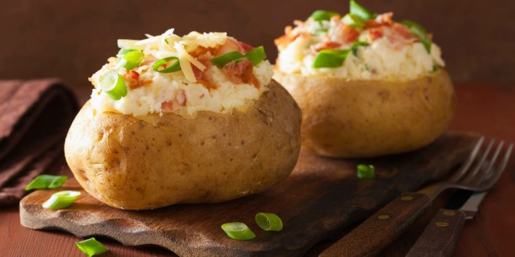 su tavolo di legno, tagliere di legno con 2 patate ripiene al forno, accanto forchetta e coltello