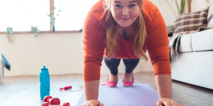 Donna bionda e sorridente in sovrappeso esegue un plank sul tappetino viola. Accanto a sé, borraccia e pesi