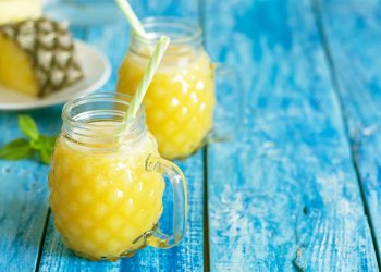tavolo di legno azzurro con 2 bicchieroni pieni di succo di ananas e bromelina e cannucce e dietro un ananas tagliato