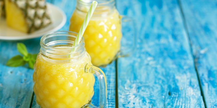 tavolo di legno azzurro con 2 bicchieroni pieni di succo di ananas e bromelina e cannucce e dietro un ananas tagliato