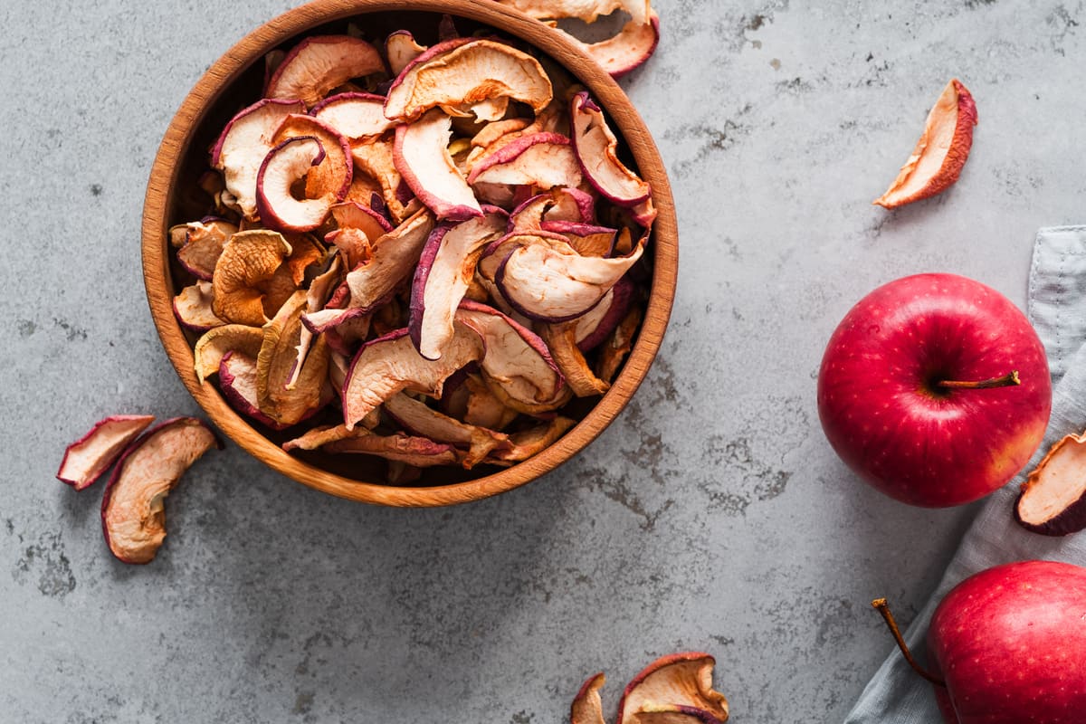 In primo piano su un tavolo di legno c'è una ciotola con delle mele essiccate, accanto dei spicchi di mela essiccati e una mela rossa.