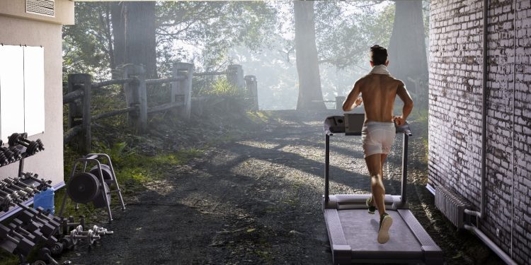 Uomo nel garage di casa sua ha creato una palestra in casa e corre sul tapis roulant sulla destra piccoli attrezzi per tonificarsi