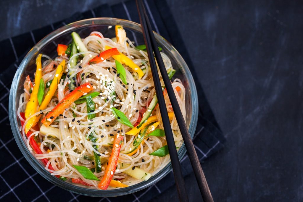 Spaghetti di soia con verdure: calorie, valori nutrizionali e come prepararli in casa 2 su sfondo nero ciotola nera con spaghetti di soia con verdure. peperoni rossi e verdi e carote. Sulla ciottola 2 bacchette cinesi per mangiarli