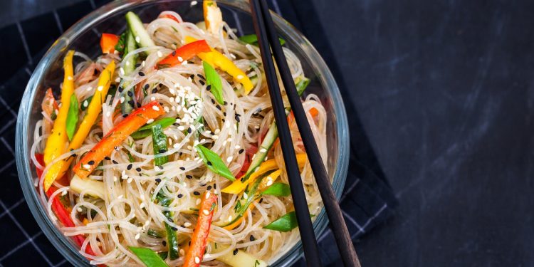 su sfondo nero ciotola nera con spaghetti di soia con verdure. peperoni rossi e verdi e carote. Sulla ciottola 2 bacchette cinesi per mangiarli
