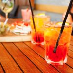 su tavolo di legno, 2 bicchieri con spritz analcolico, ghiaccio, limone e cannucce accanto un tagliere di legno con olive, pizette e tramezzini, accanto busto di una dona con maglietta nera che sta bere aperitivo