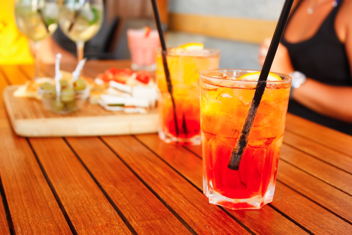 su tavolo di legno, 2 bicchieri con spritz analcolico, ghiaccio, limone e cannucce accanto un tagliere di legno con olive, pizette e tramezzini, accanto busto di una dona con maglietta nera che sta bere aperitivo