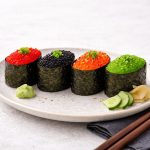 In primo piano su un tavolo chiaro c'è un piatto con dei gunkanmaki di lompo, delle fettine di cetriolo, zenzero e salsa wasabi. Sulla destra su un tovagliolo grigio ci sono due bacchette in legno.