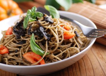 Primo piano di un piatto bianco con spaghetti al nero di seppia, pomodori e prezzemolo con una forchetta sul bordo. In secondo piano un pomodoro