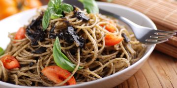 Primo piano di un piatto bianco con spaghetti al nero di seppia, pomodori e prezzemolo con una forchetta sul bordo. In secondo piano un pomodoro
