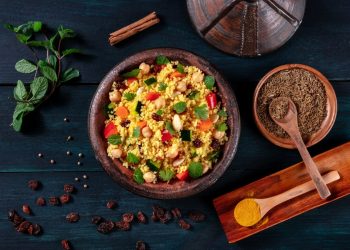 una ciotola piena di cous cous accanto a erbe aromatiche e utensili di legno